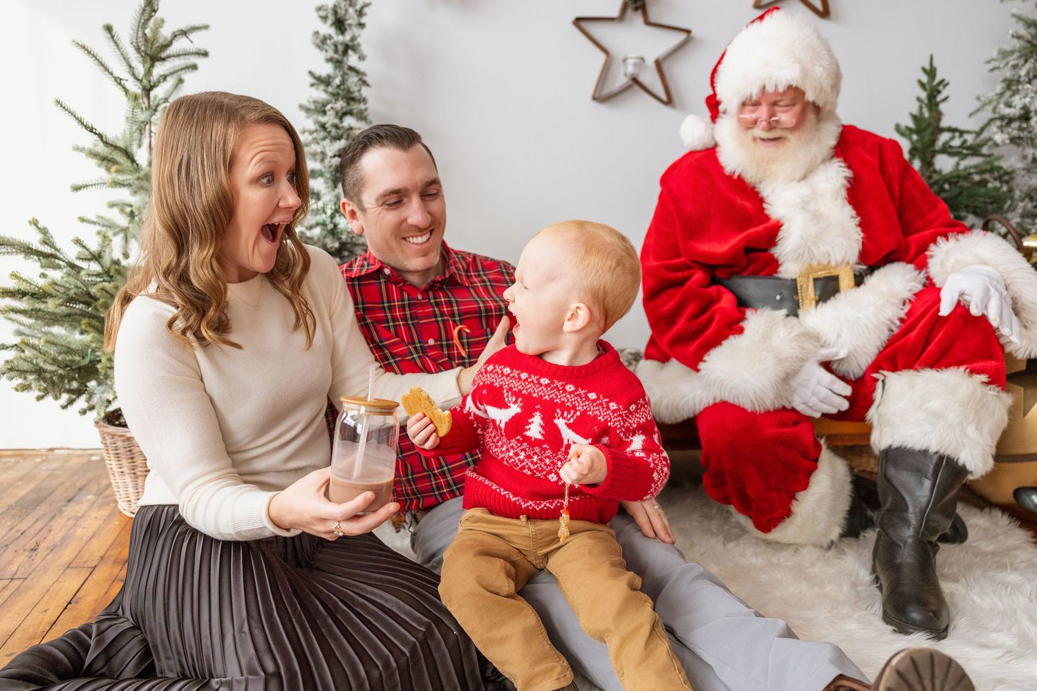 Young family with baby in red reindeer sweater enjoys cheerful moment with Santa at Christmas.