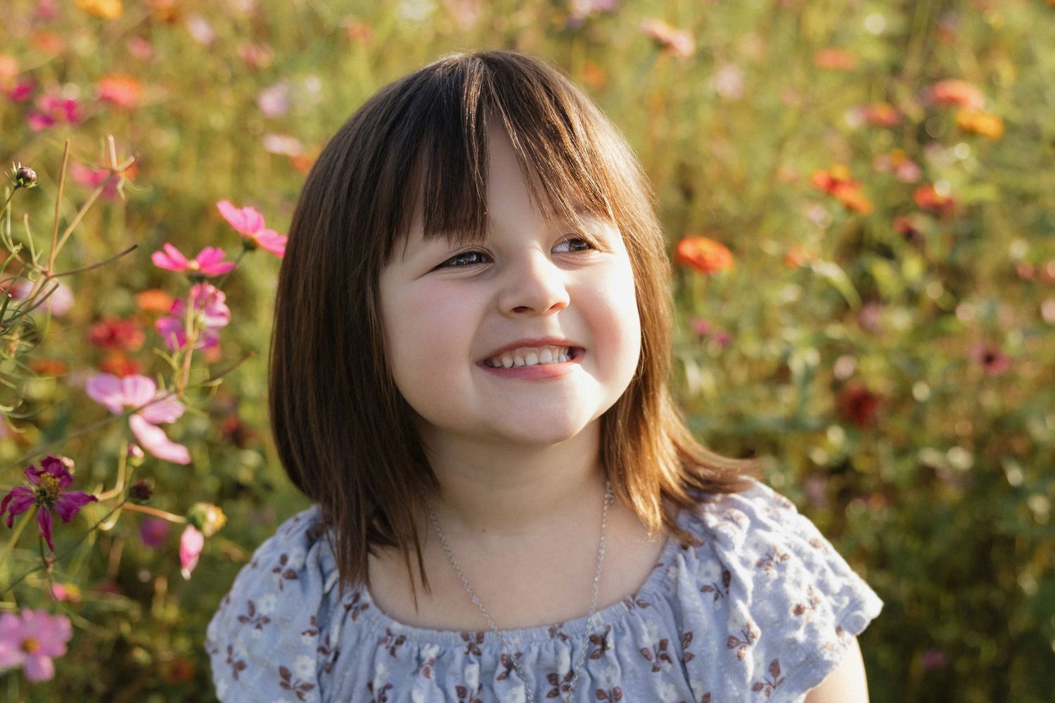 A cheerful young girl with bangs smiles brightly in a sunlit flower field with pink and orange blooms.