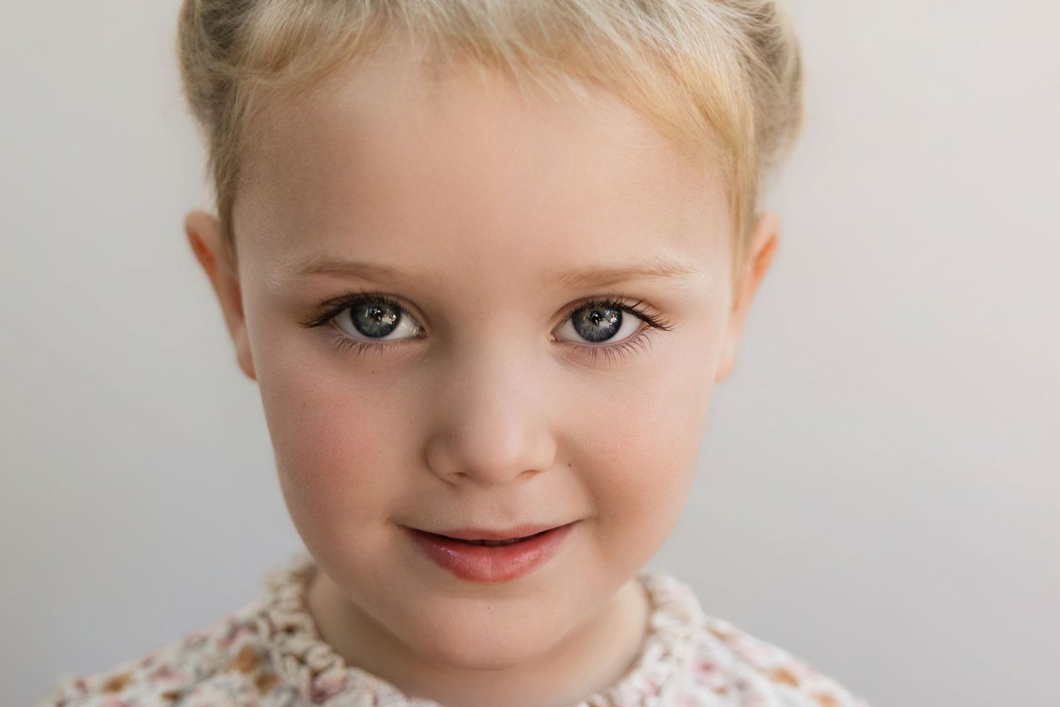 Close-up portrait of a young blonde girl with green eyes and a gentle smile against a white background.