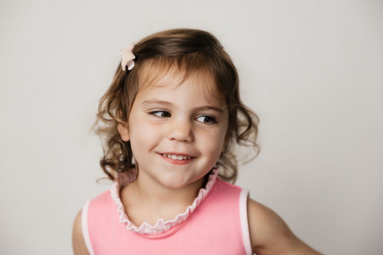 Smiling young girl with curly brown hair wearing a pink sleeveless dress and pearl necklace against a white background.