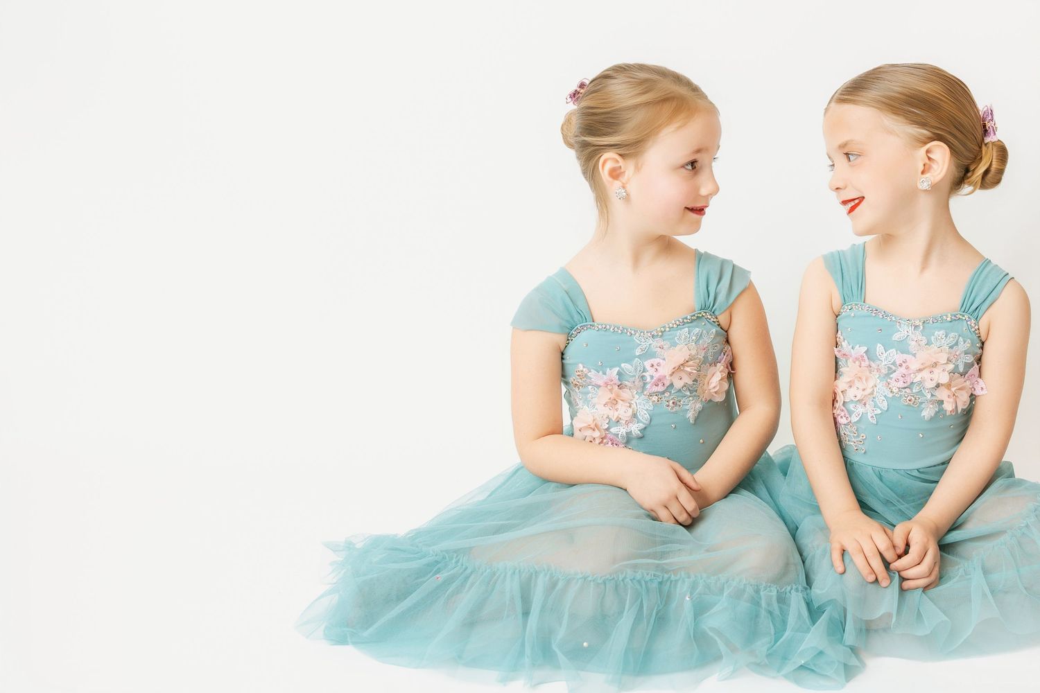 Two young girls in matching teal floral dresses sitting together and smiling at each other.