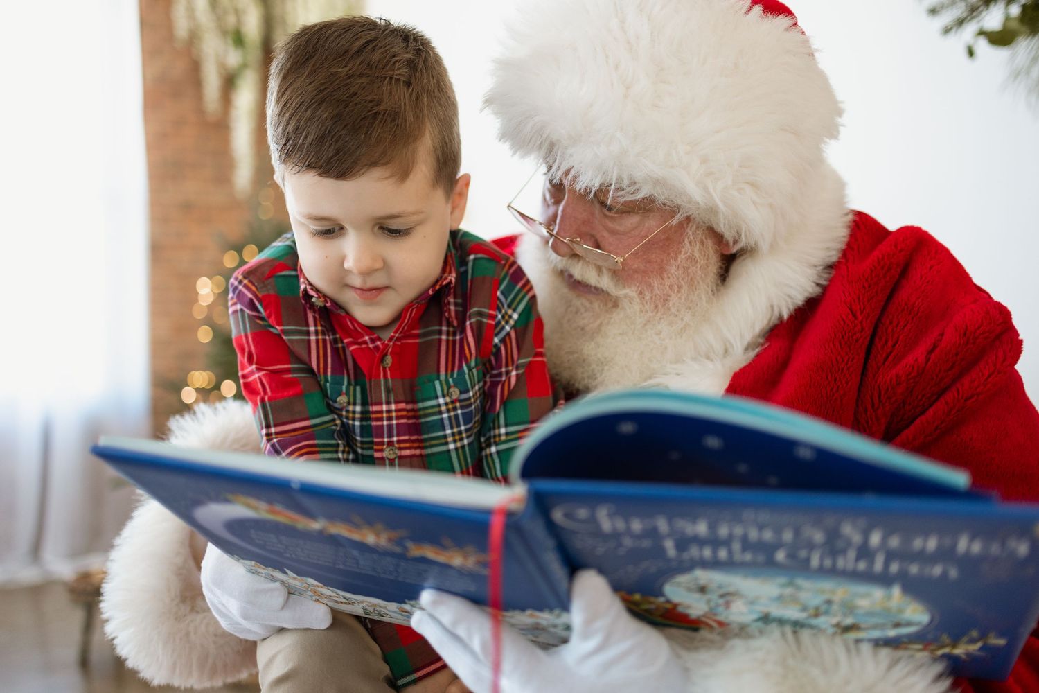 Santa Claus reads a Christmas storybook with a young boy in plaid pajamas during the holiday season.
