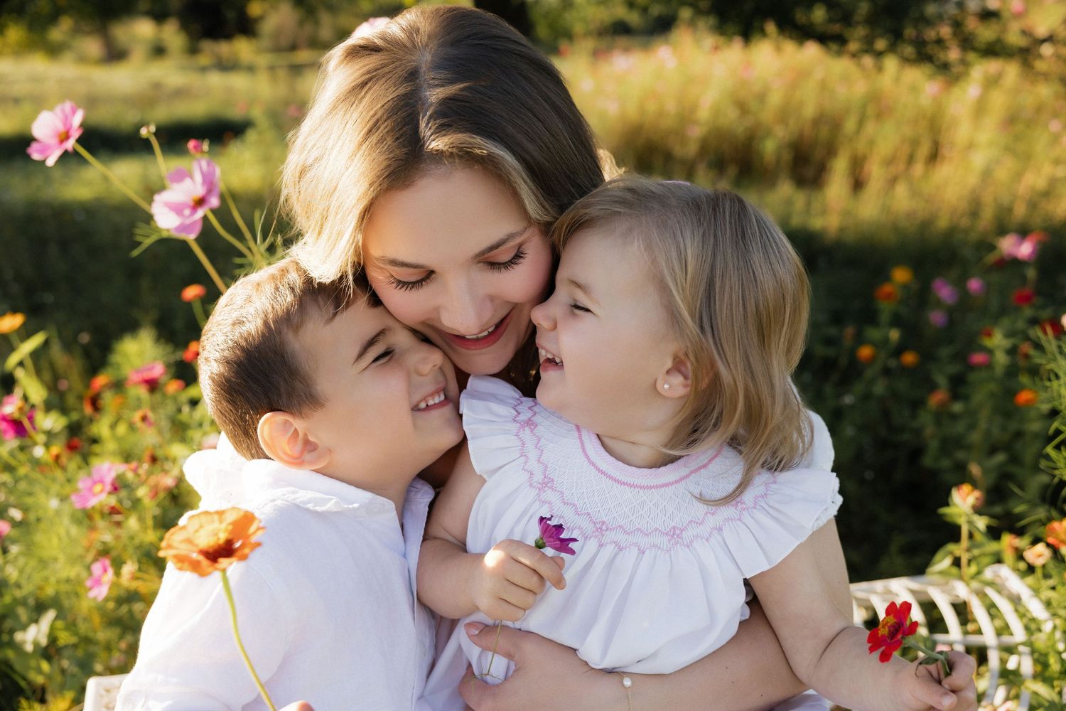 A mother hugs her two young children in a sunny flower garden, sharing a joyful and loving family moment.