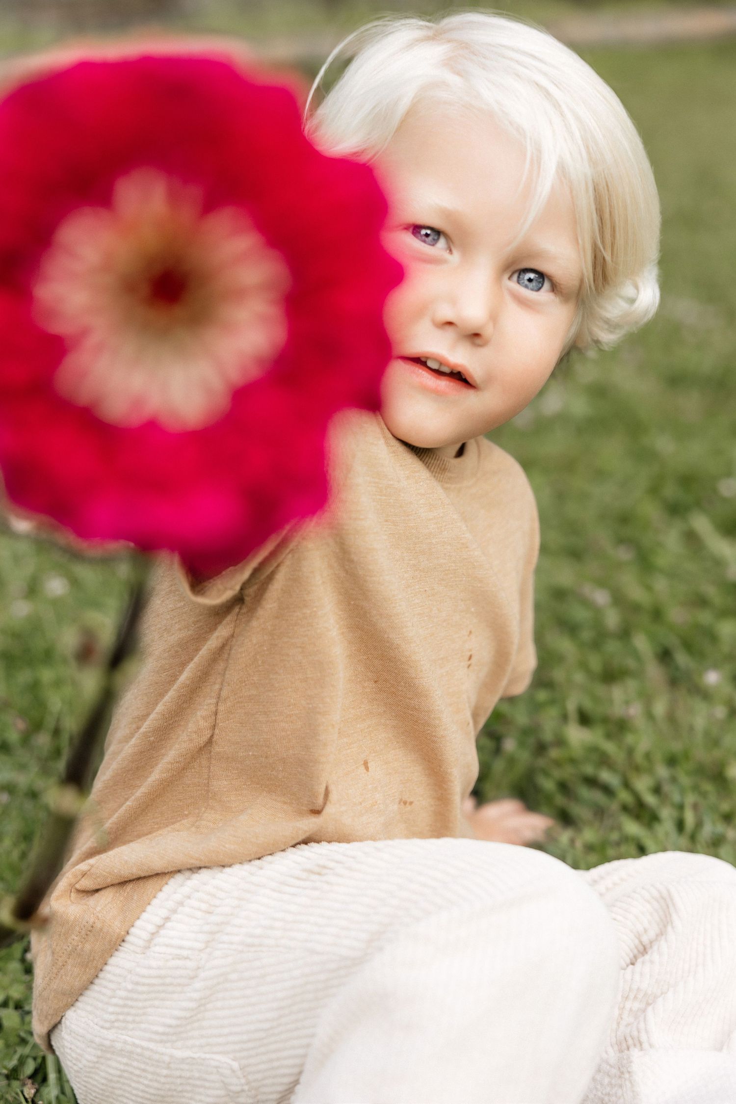 Blonde toddler with blue eyes holding a red flower outdoors on green grass in a beige linen outfit.