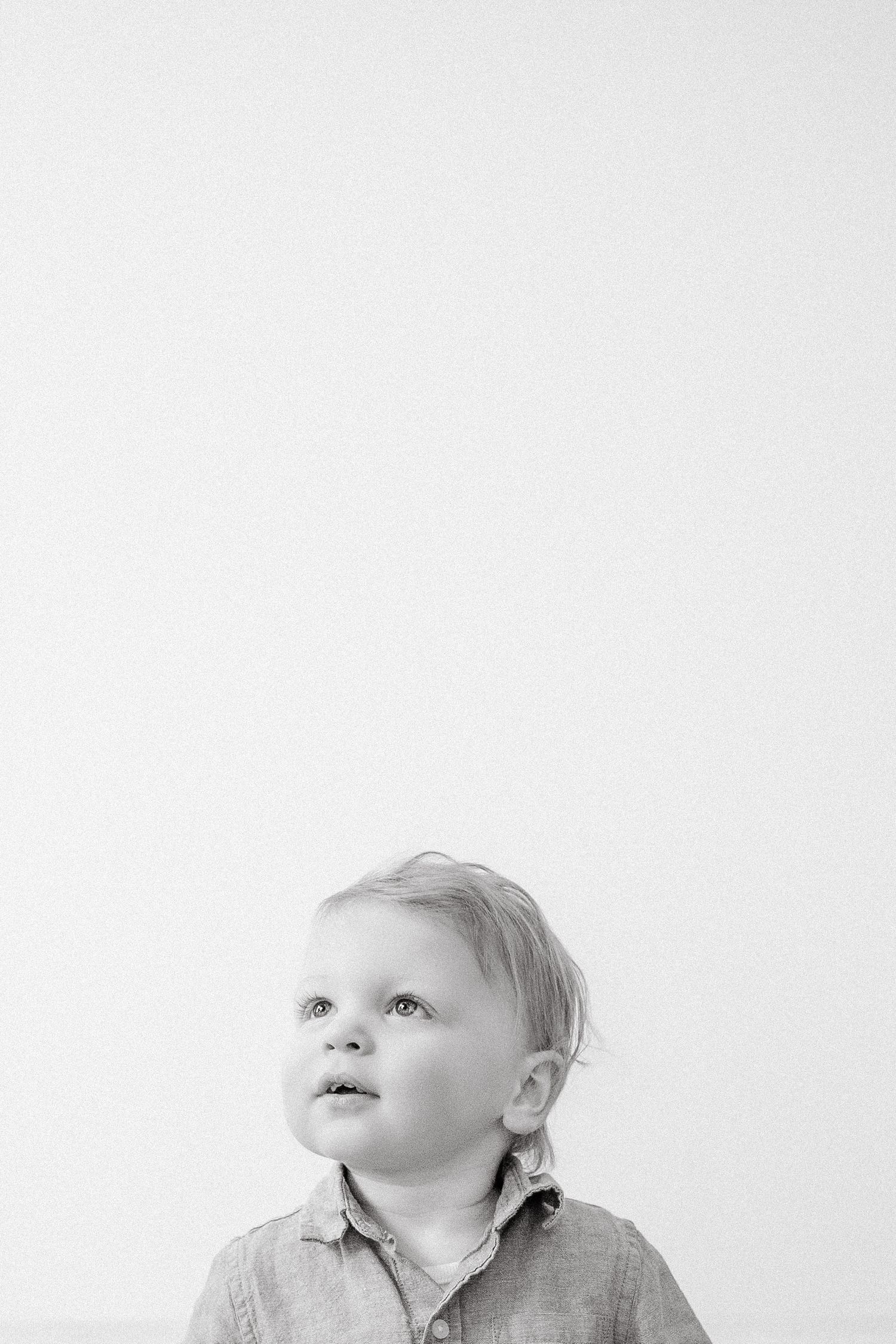 A black and white portrait of a curious toddler in a linen shirt looking upward against a white wall.