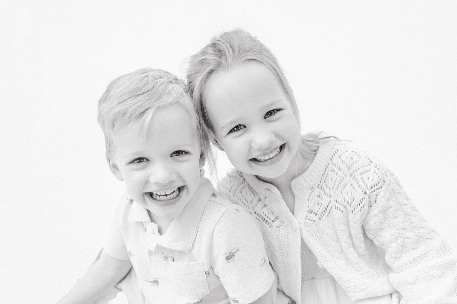 Two smiling children posing together in a bright black and white sibling portrait photo.