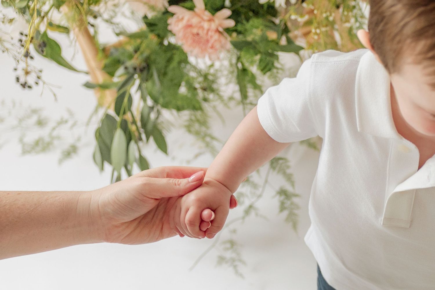 Mother gently holds a baby's tiny hand in front of a soft floral arrangement with pink and green flowers.