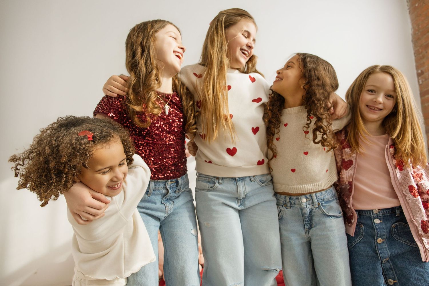 Five diverse girls laughing and hugging in heart-print sweaters and jeans.