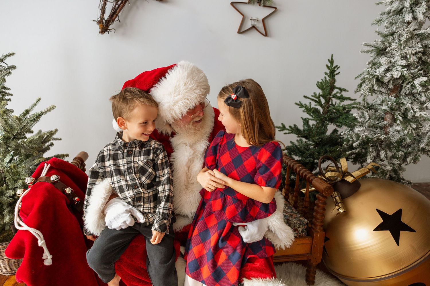 Santa Claus smiles with two children beside Christmas trees and a large golden bell decoration.