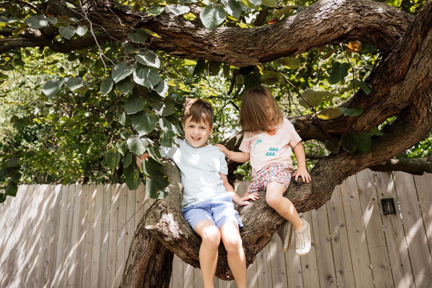 Two children sitting together on a large tree branch in a backyard, smiling and enjoying outdoor play.