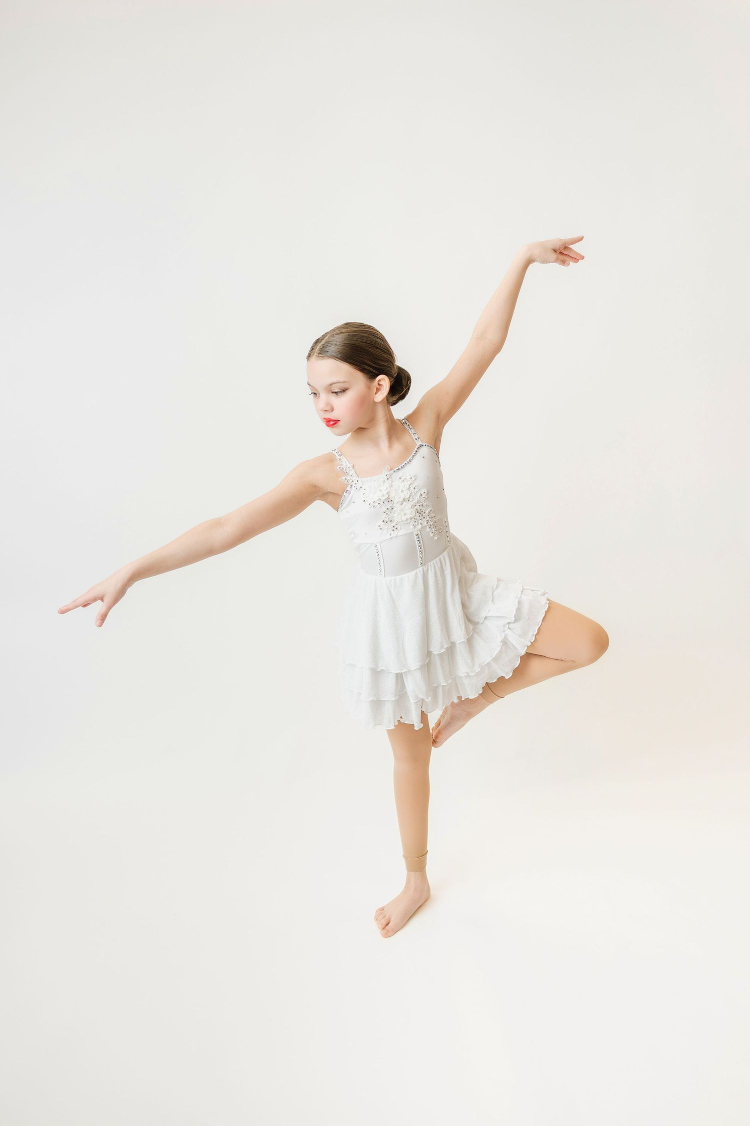 Young girl in white layered dance dress performing a graceful one-legged balance pose with arms extended.
