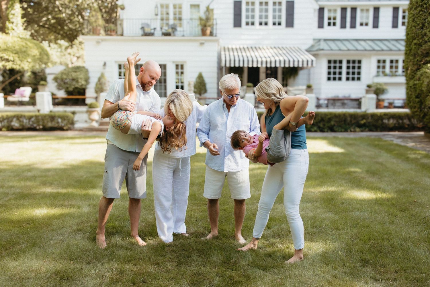 Multi-generational family playing together barefoot on green lawn outside elegant white home.