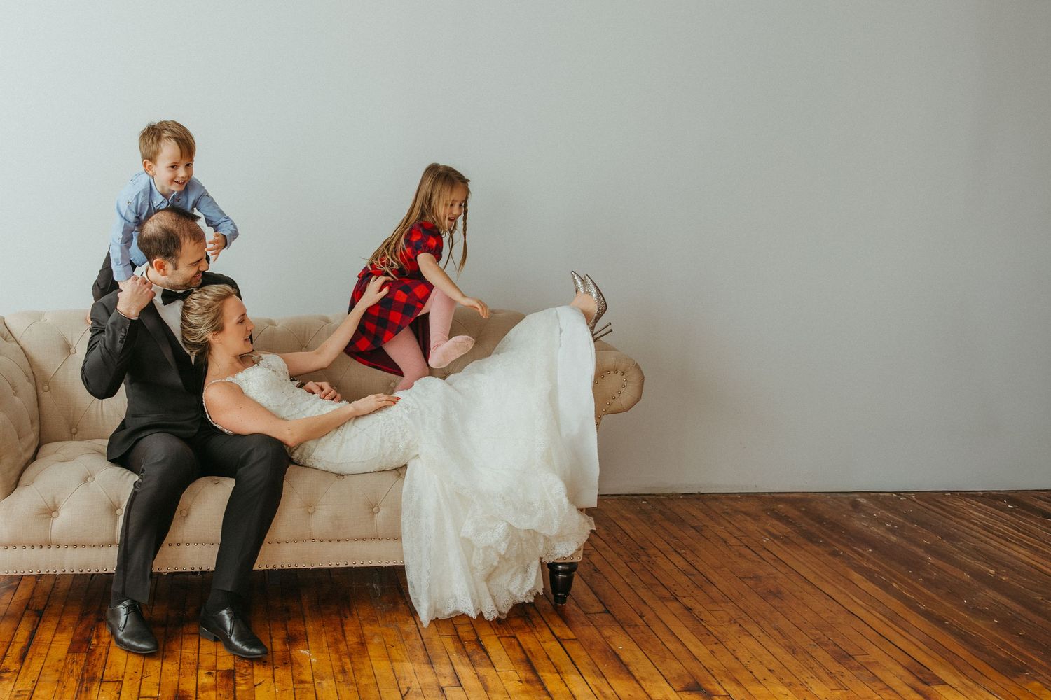 Bride and groom celebrating anniversary. They sitting on a couch with their two playful young children climbing on them.
