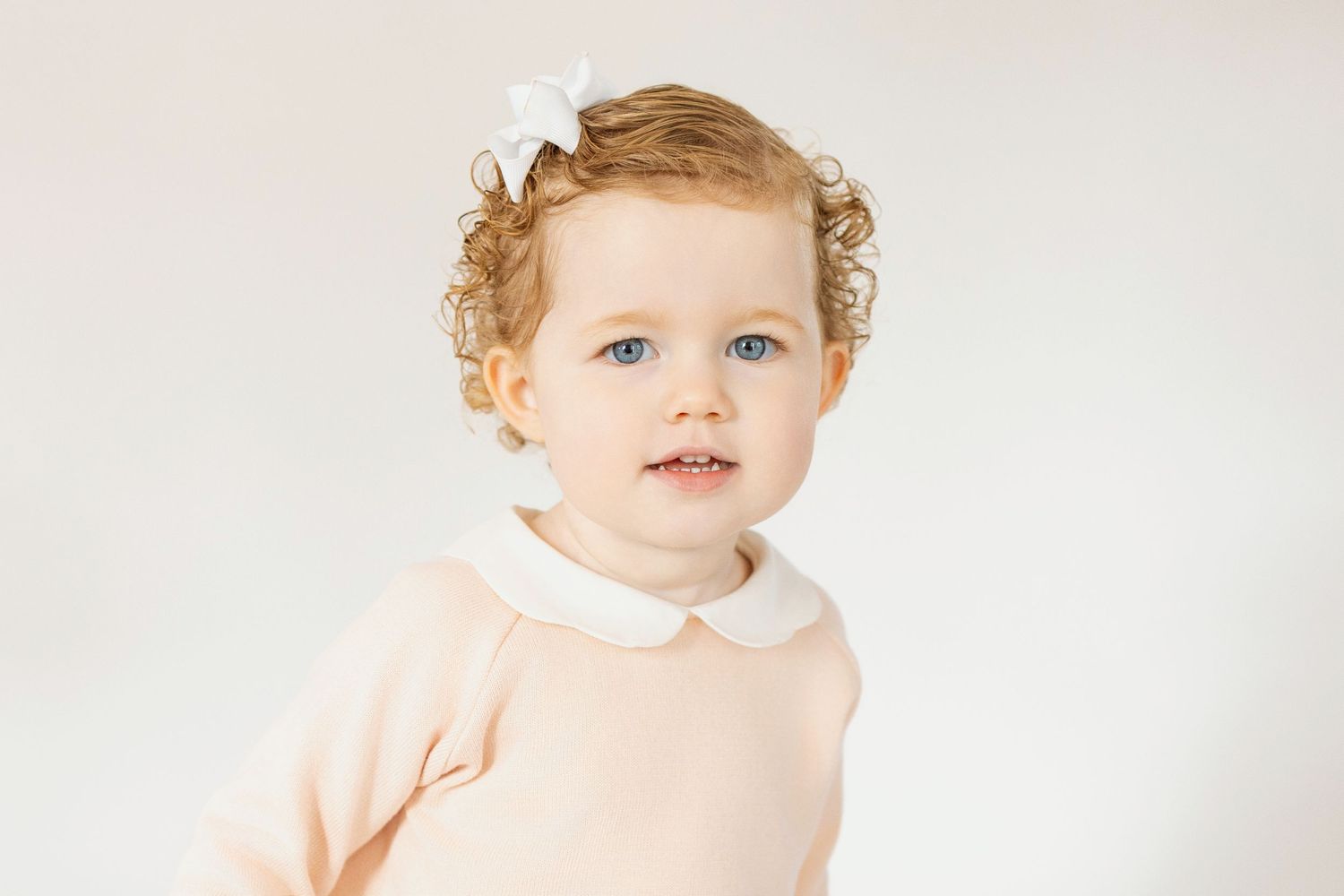 Adorable toddler girl with curly red hair and blue eyes wearing a pink dress with white collar and bow.