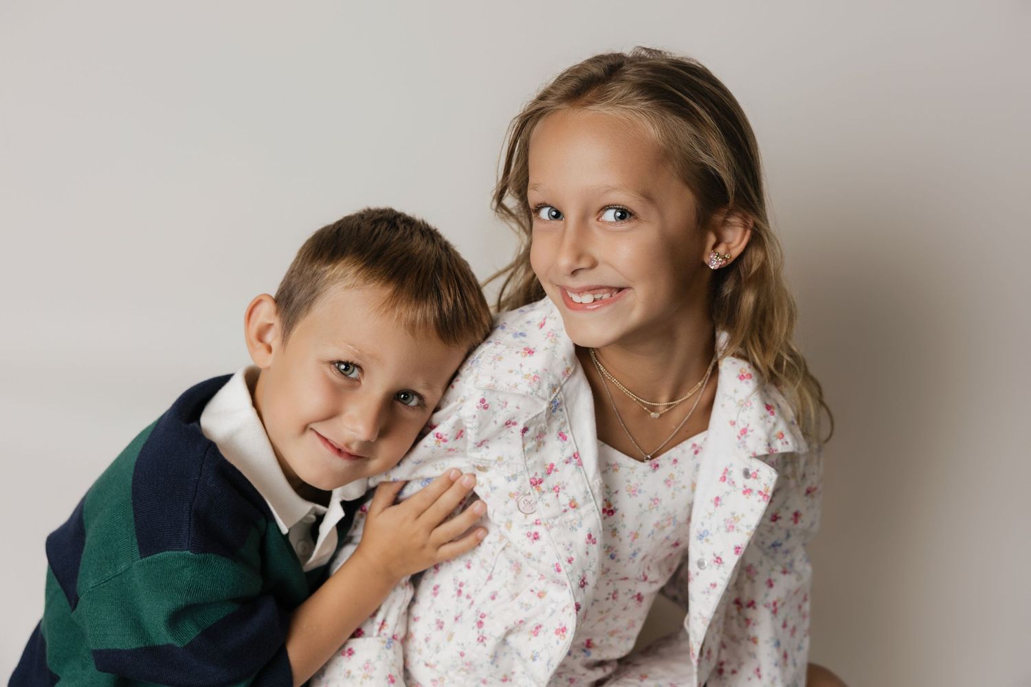 Two smiling children, a young boy and older girl, pose together for a portrait photo against a light background.