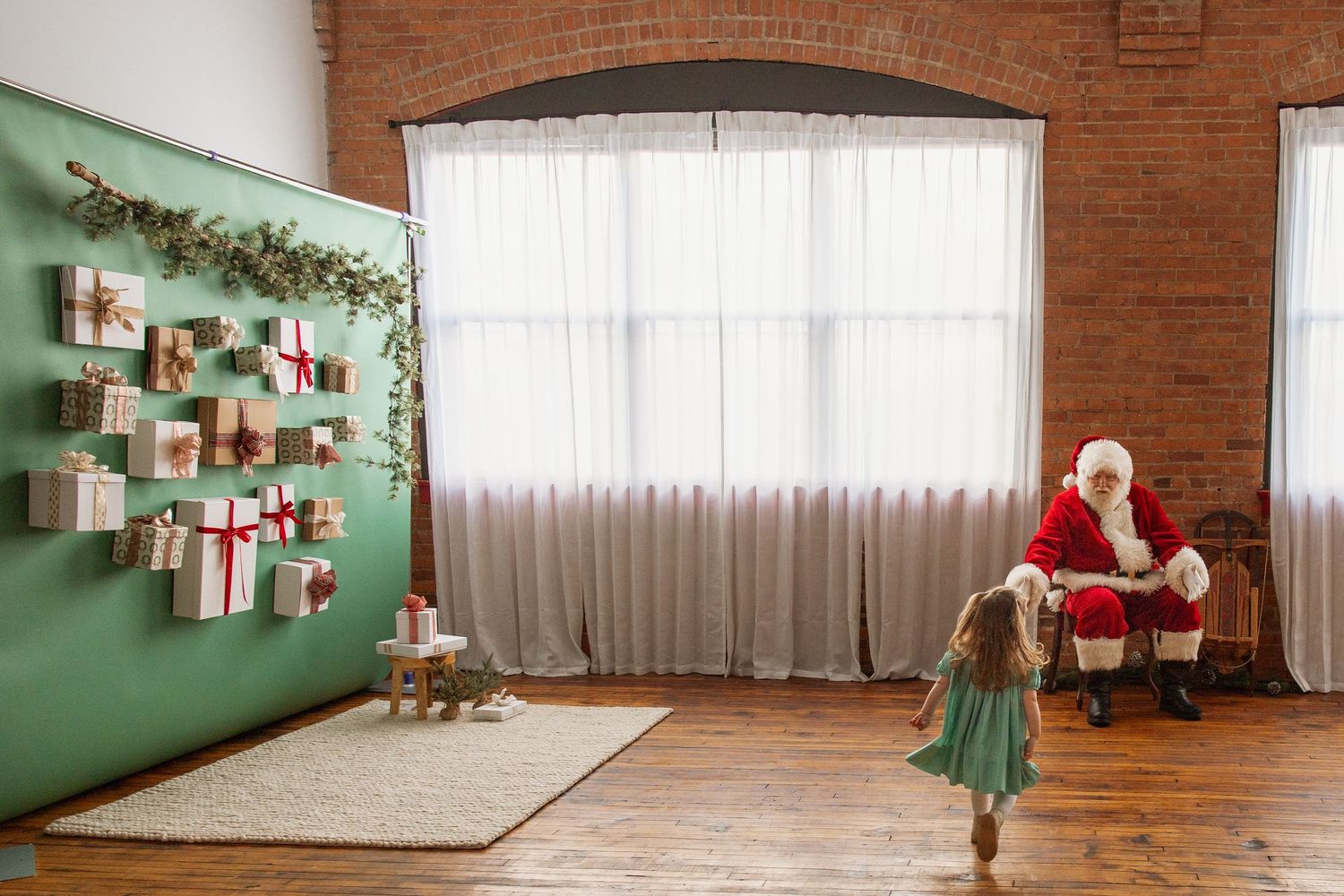 Young girl runs toward Santa Claus seated in festive room with wrapped gifts on green wall.