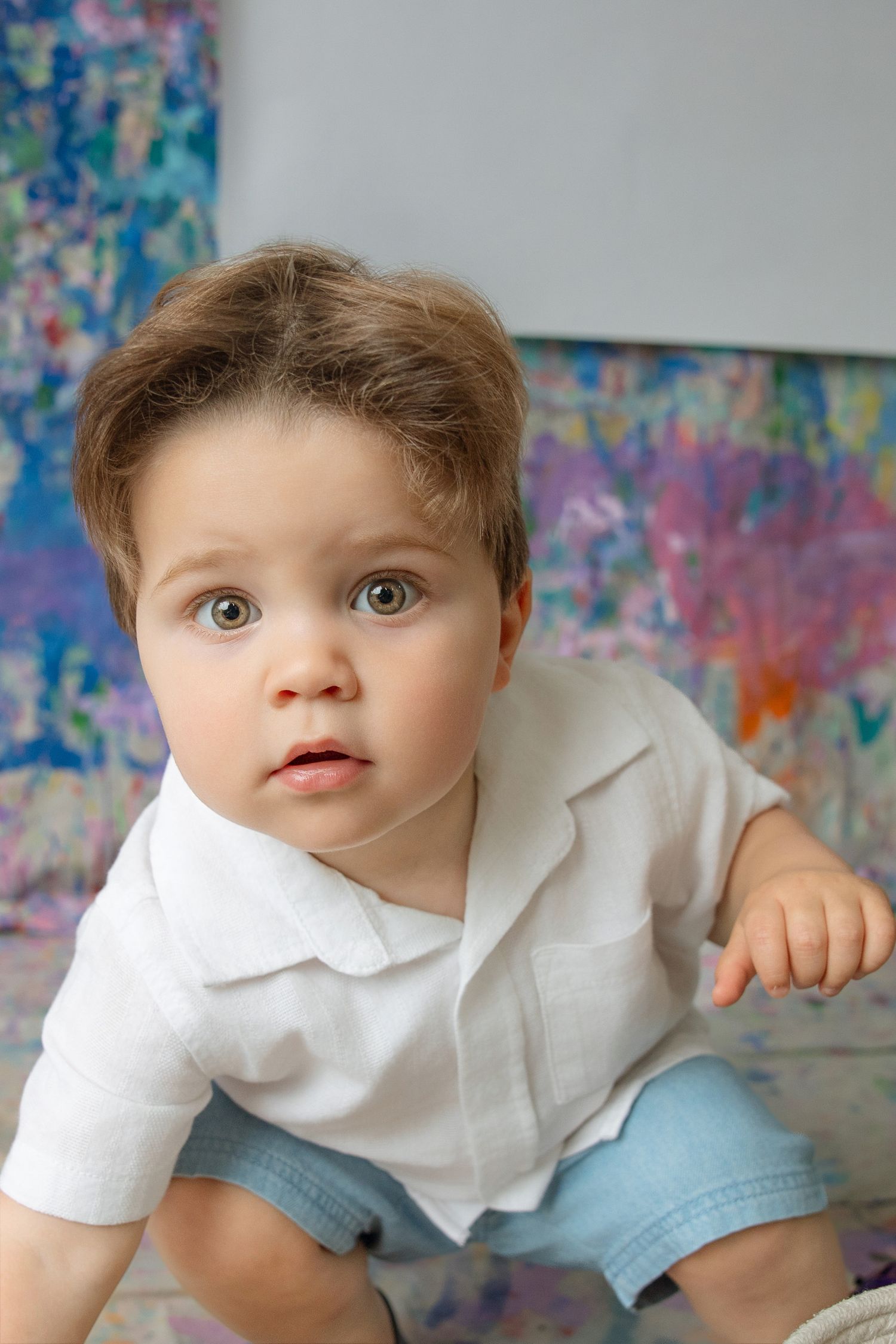 Adorable toddler boy in white shirt and blue shorts looking curious against colorful abstract painting background.