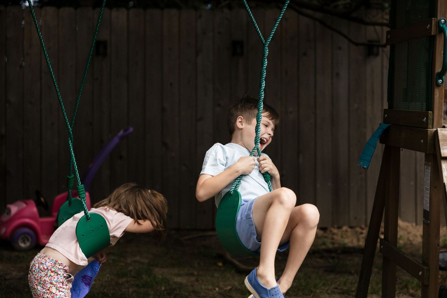 Two children playing on a green rope swing set in a backyard, with a wooden fence in the background.