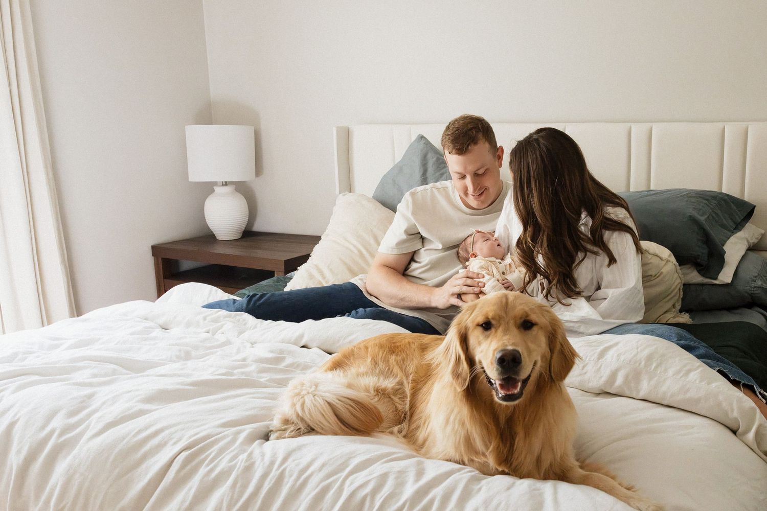 Couple with newborn relaxing on a white bed with a golden retriever dog in a bright, cozy bedroom with neutral decor.