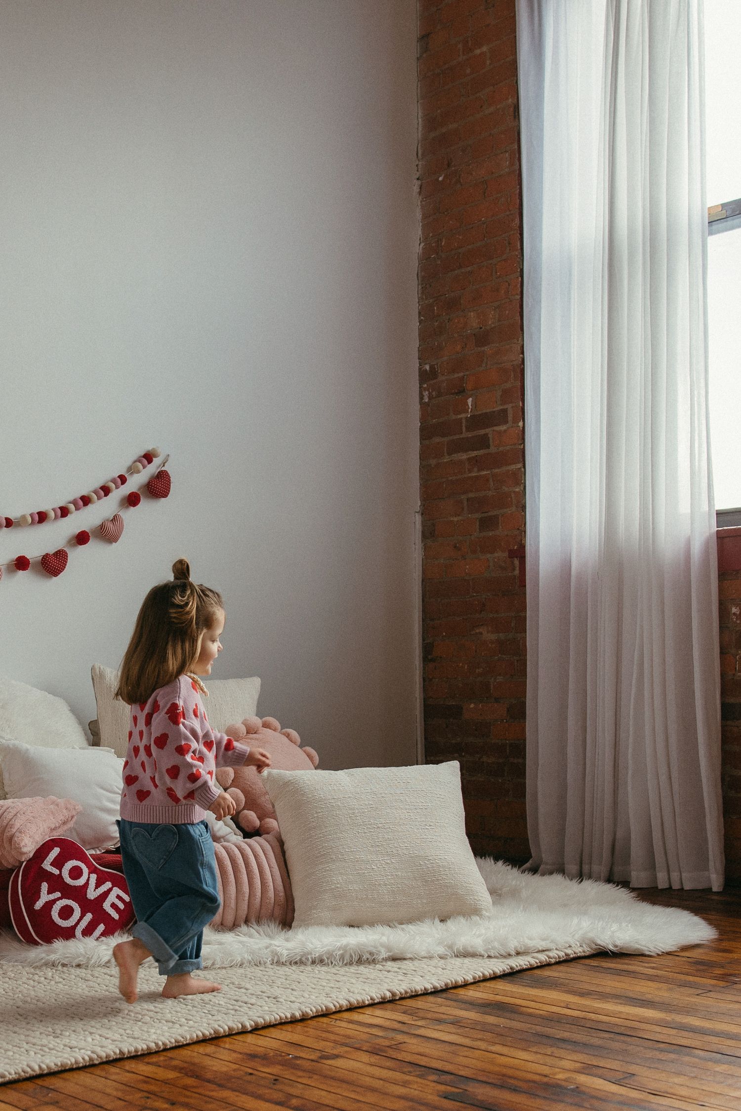Young girl in pink sweater playing near pillows in decorated bedroom with brick wall.