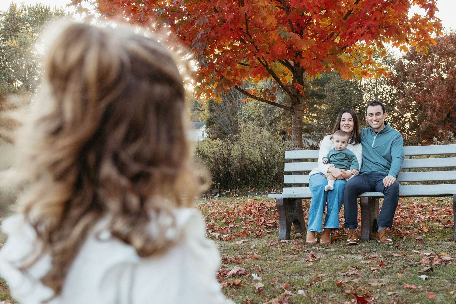 A couple sits on a white bench holding a baby as a child walks toward them in autumn.