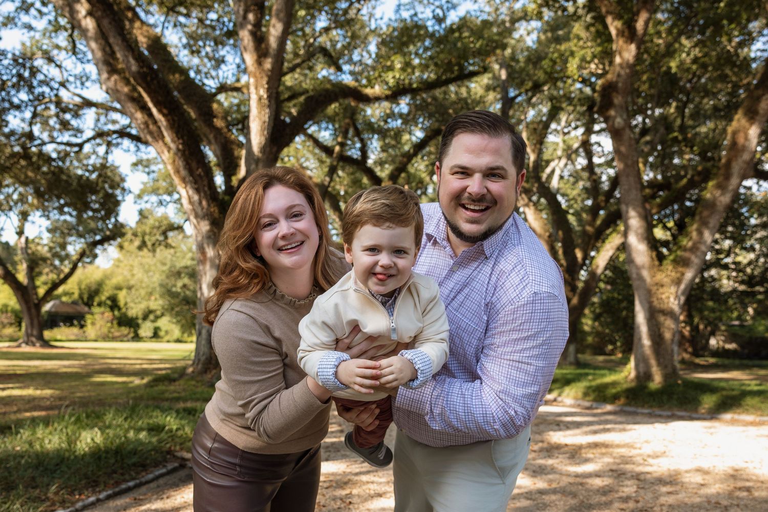 Smiling family of three posing together under large oak trees outdoors.