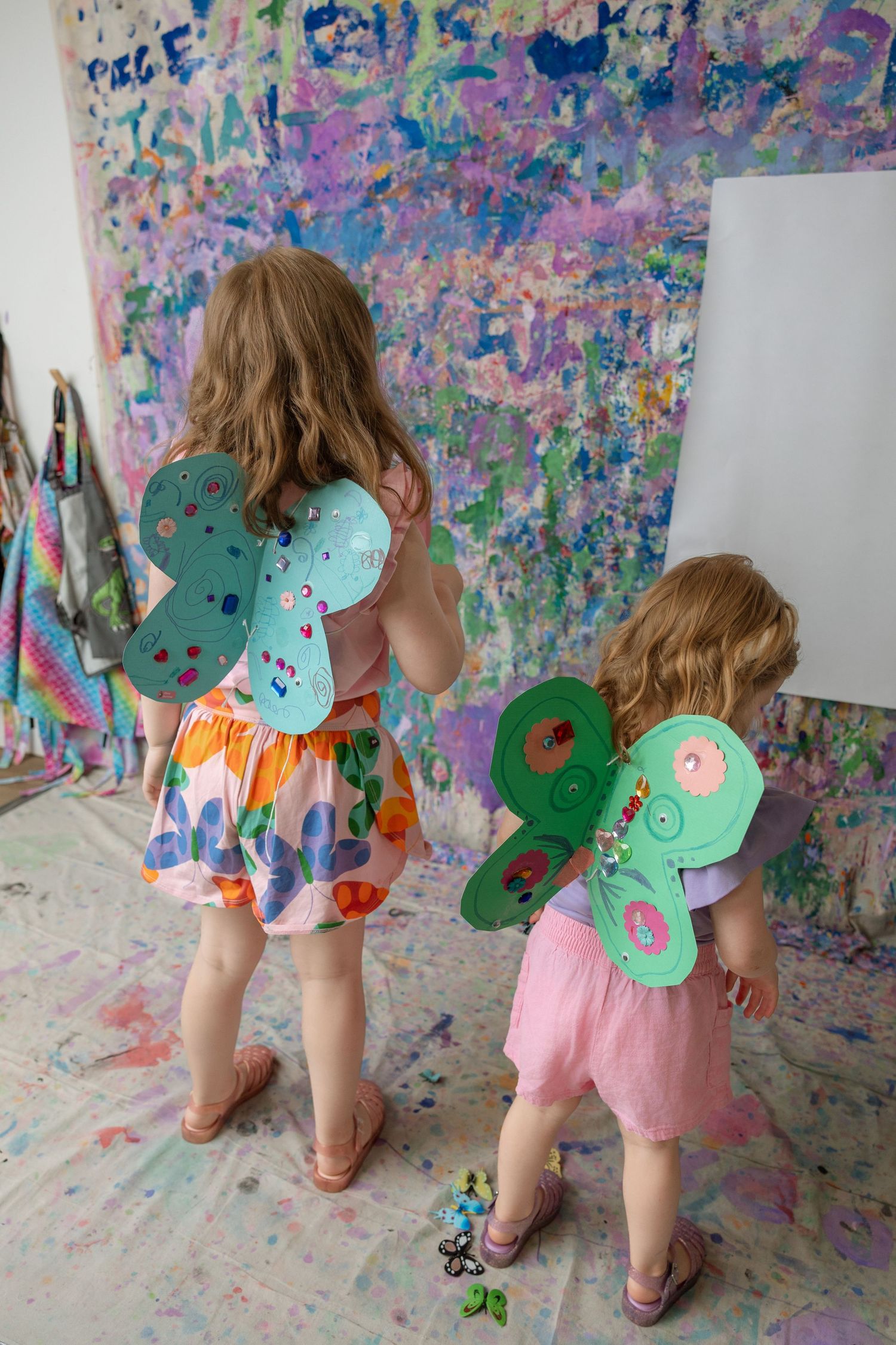 Two young girls wearing colorful butterfly wings stand before a paint-splattered wall in an art studio.