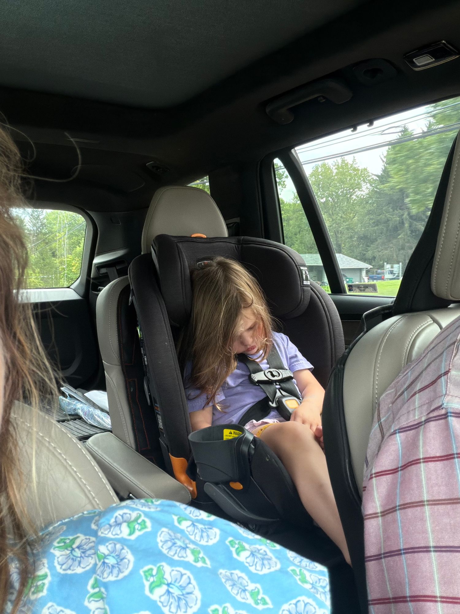Young girl secured in a black car seat in the back of an SUV, looking down while traveling.