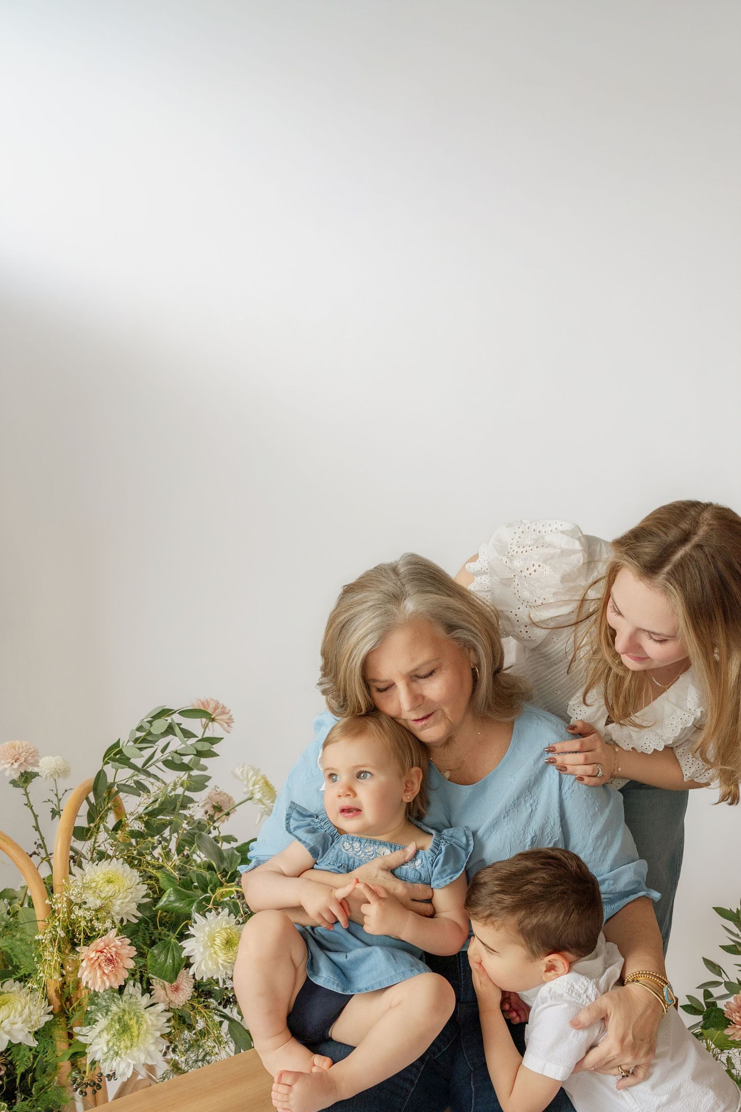A grandmother sits with her daughter and two grandchildren surrounded by fresh flowers in a bright, airy white studio setting.