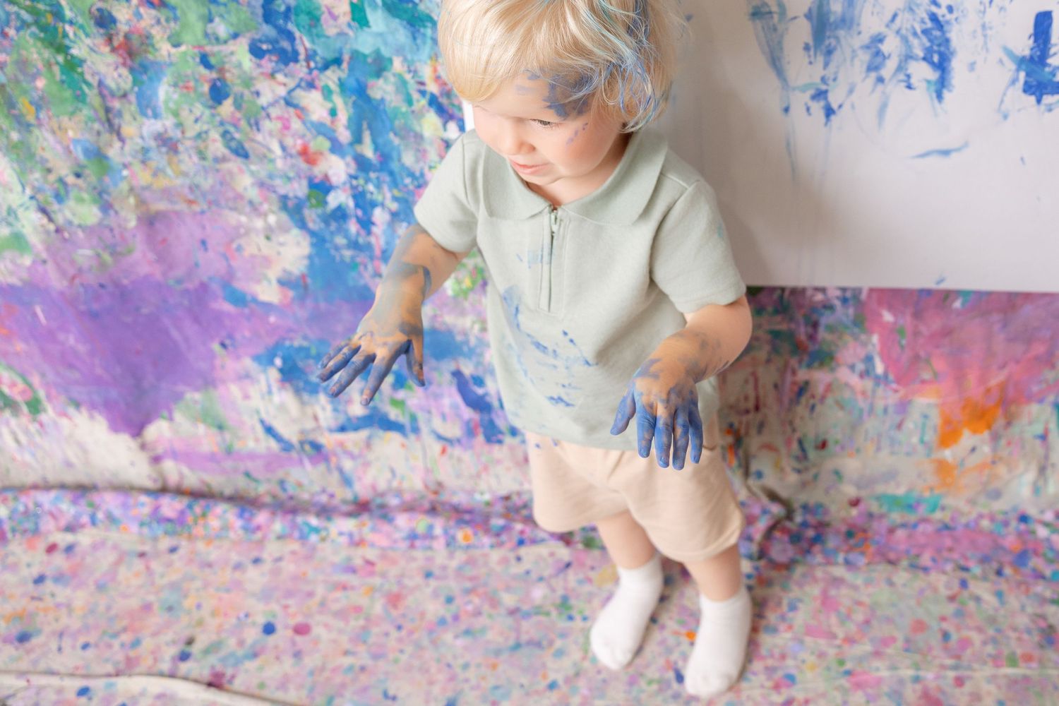 Toddler with blue paint-covered hands standing in front of a colorful painted wall and floor during a creative art session.