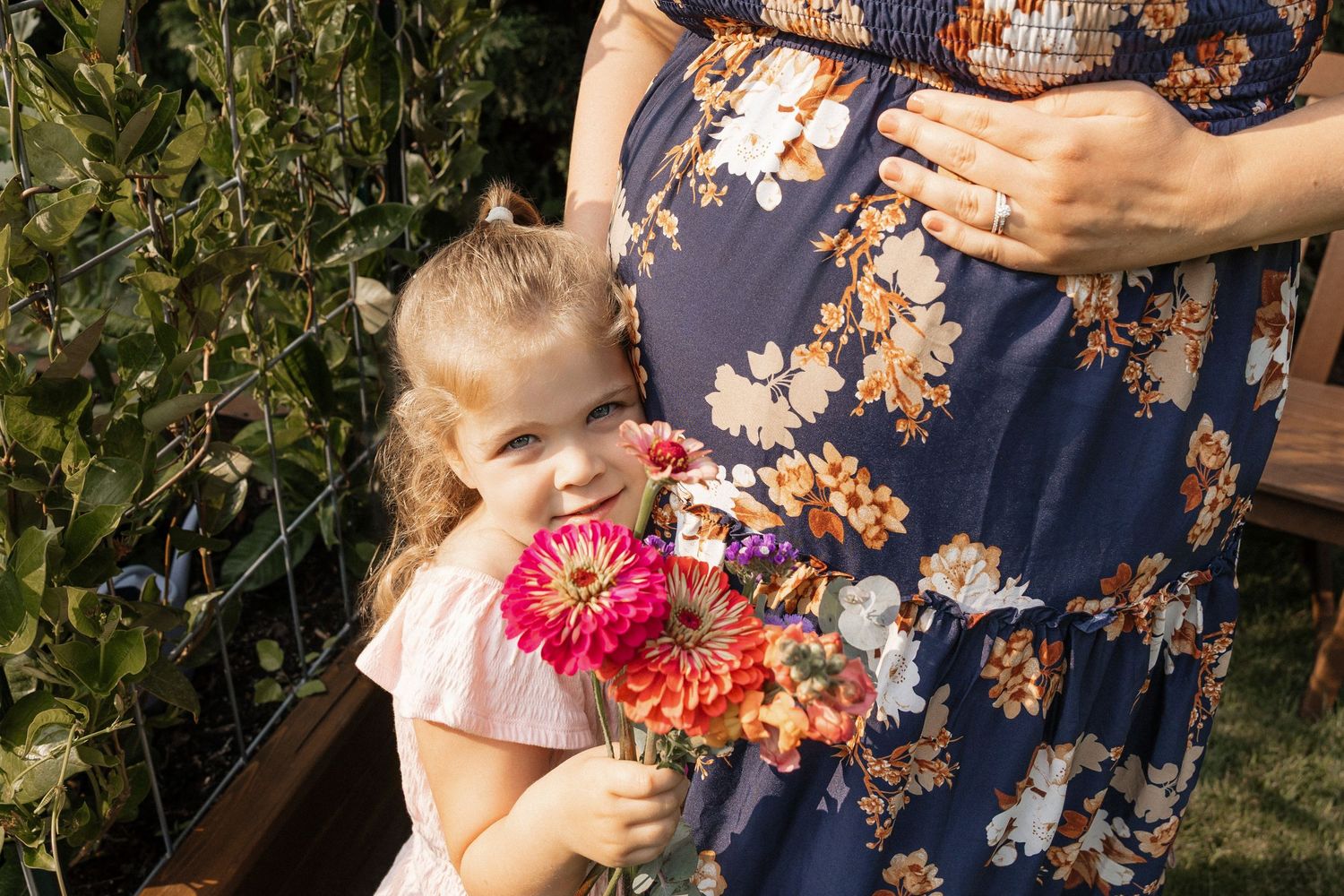A young girl holds colorful zinnias beside a pregnant woman in a floral dress outdoors.
