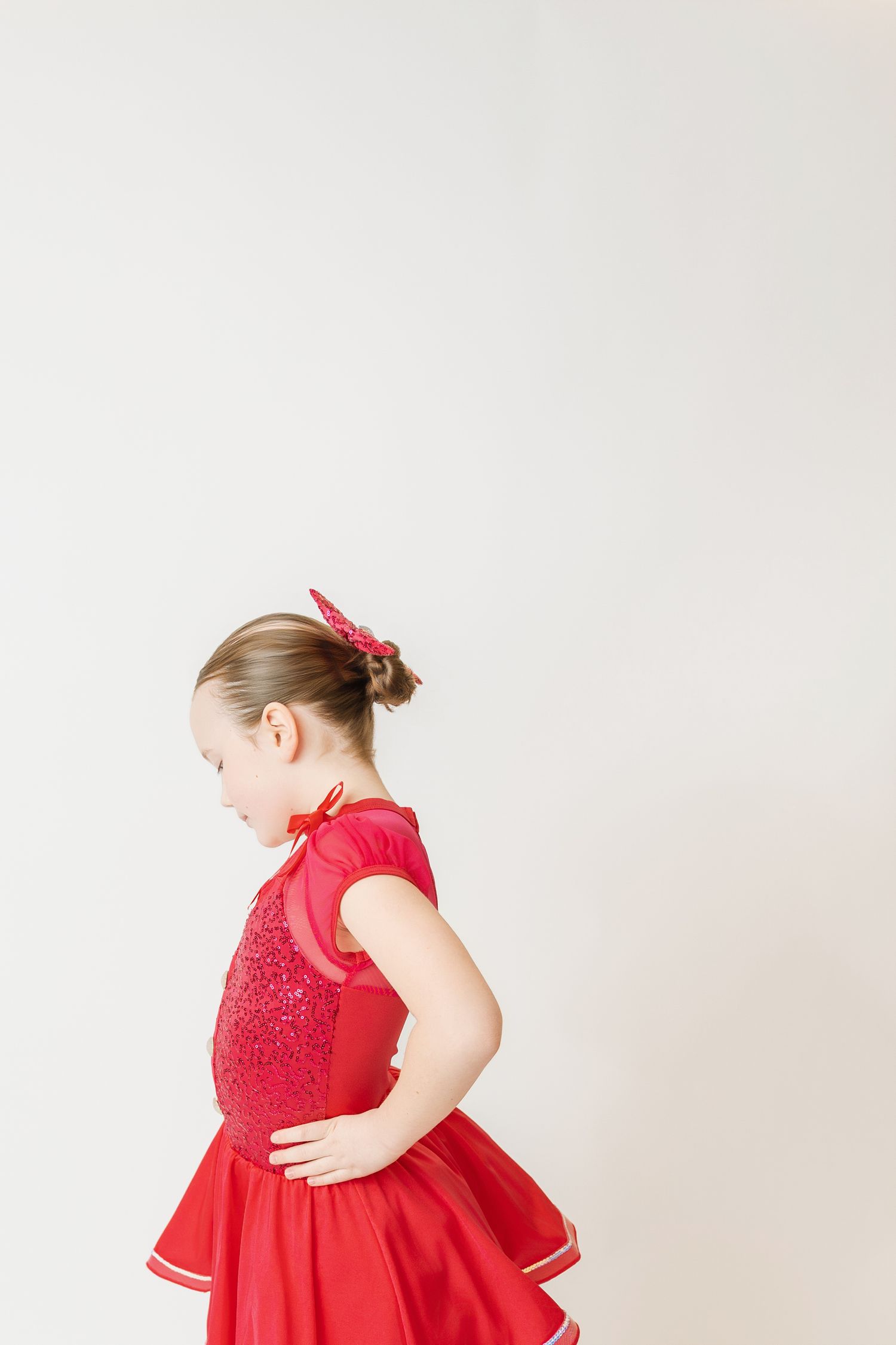 Young girl in red sequin dance costume with matching bow looking down gracefully against white background.