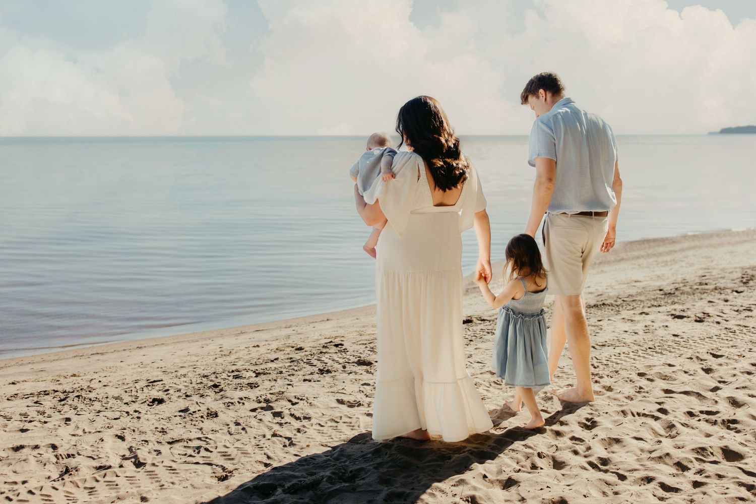 Family of four walking hand-in-hand on a sandy beach, with calm ocean waters and a cloudy sky in the background.