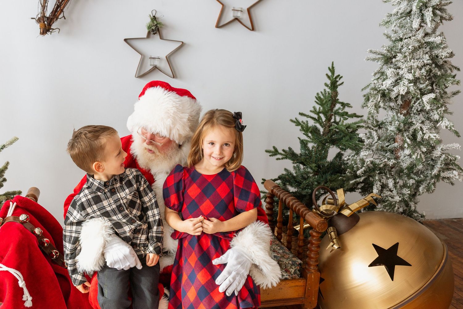 Two children pose with Santa Claus beside a decorated Christmas tree with star ornaments and a gold bell.