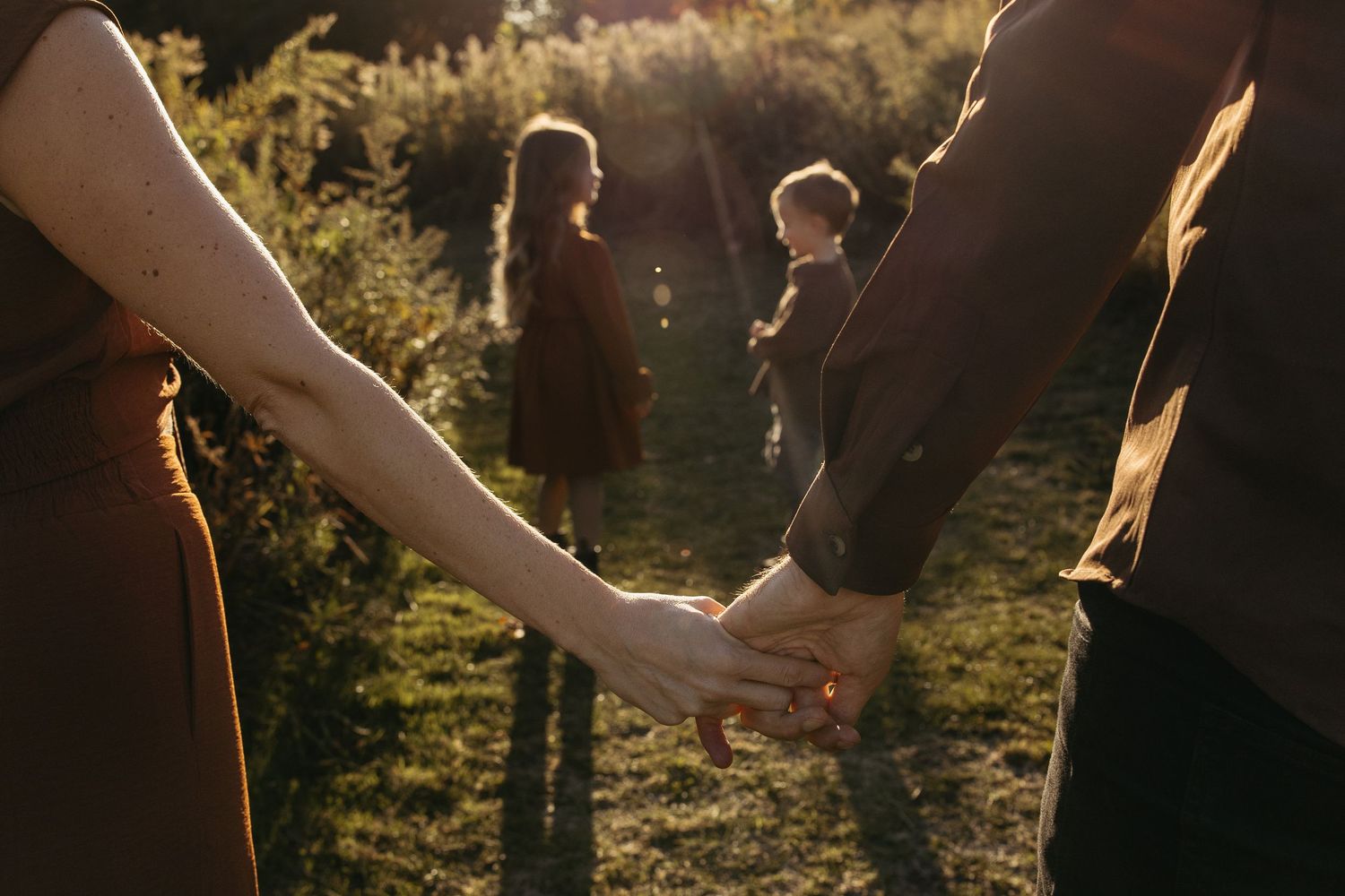 Couple holding hands in warm sunlight while two children play in green meadow background.