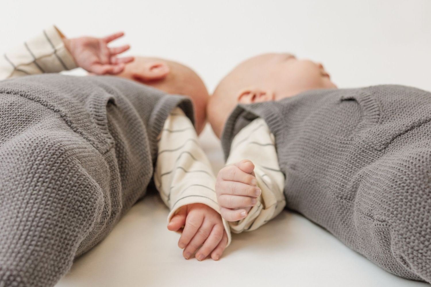 Two baby twins in matching gray knit outfits lie side by side, their tiny hands nearly touching on a white surface.