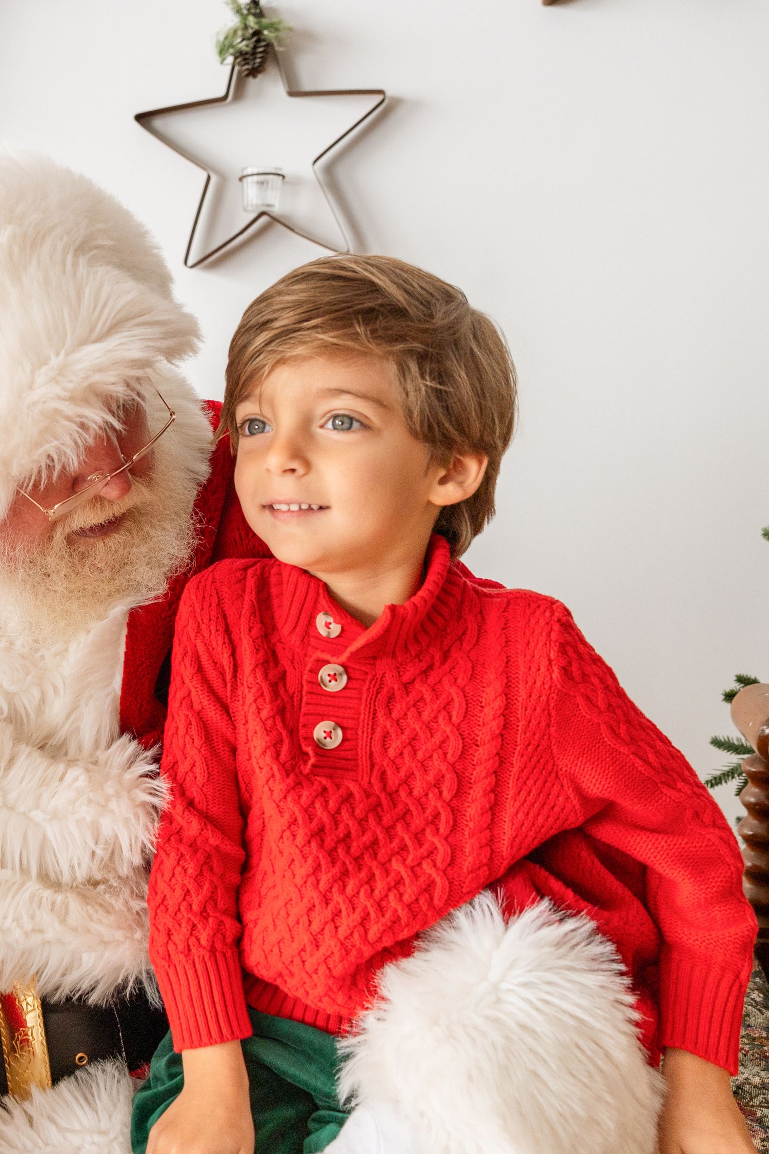 A blue-eyed boy in a red cable-knit sweater leans against Santa during a Christmas photo session.