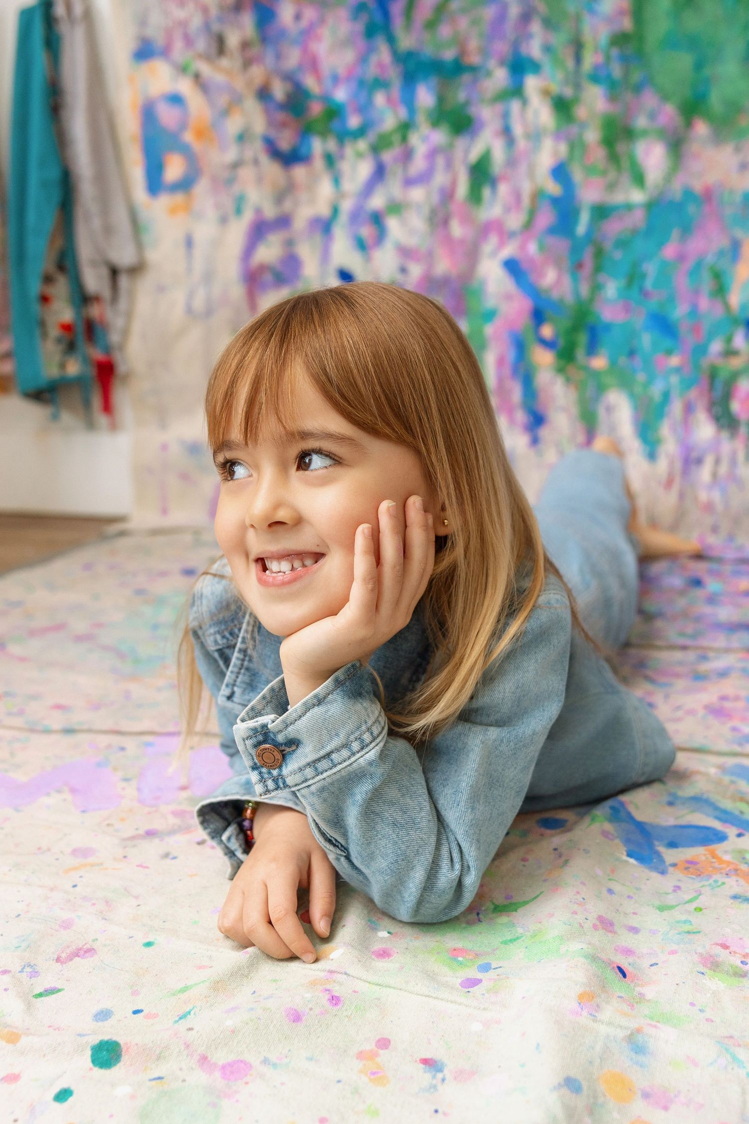 A smiling girl in denim lies on a paint-splattered floor before a colorful abstract mural.