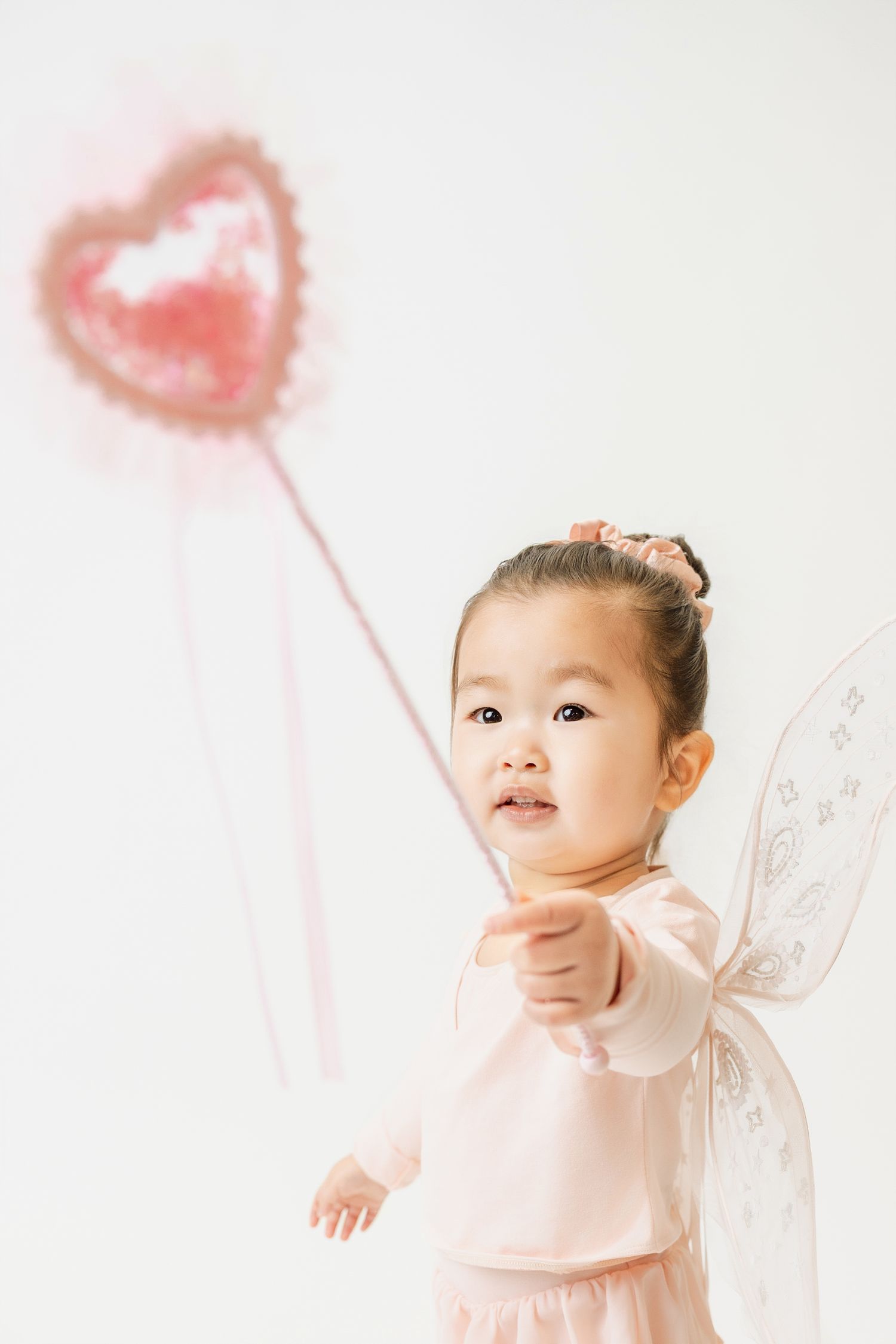 Toddler girl in pink fairy costume with wings holding a heart-shaped wand against white background.