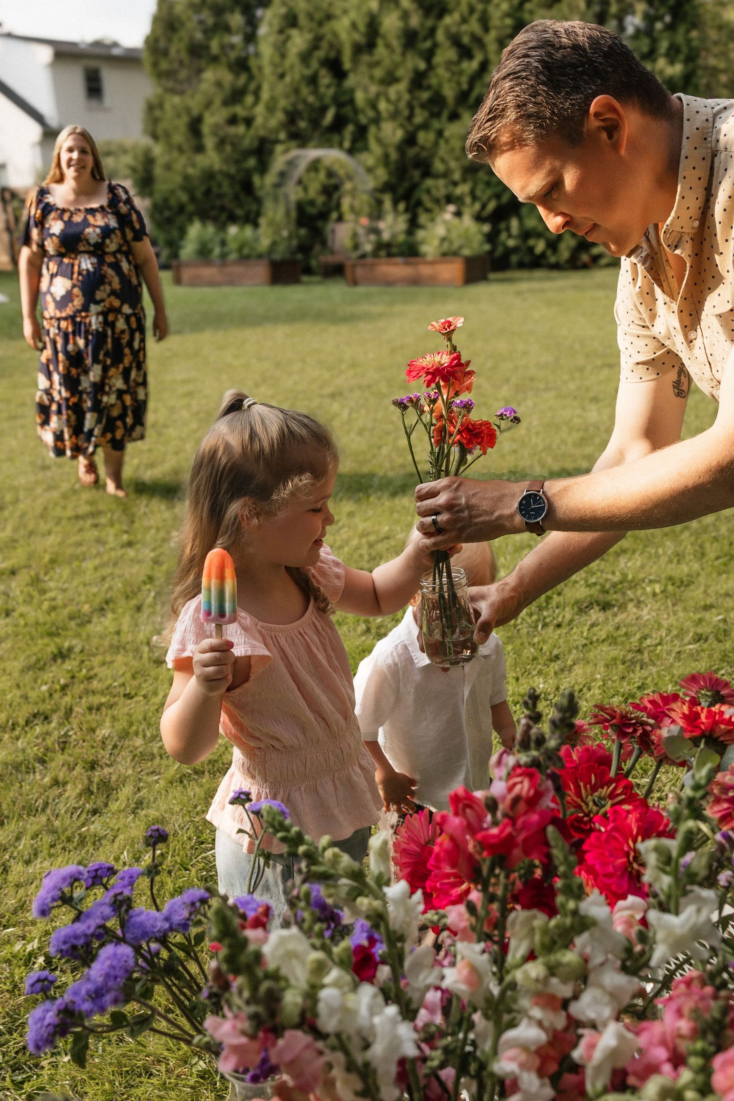Father and daughter picking colorful flowers in a garden while mother walks toward them on a sunny day.
