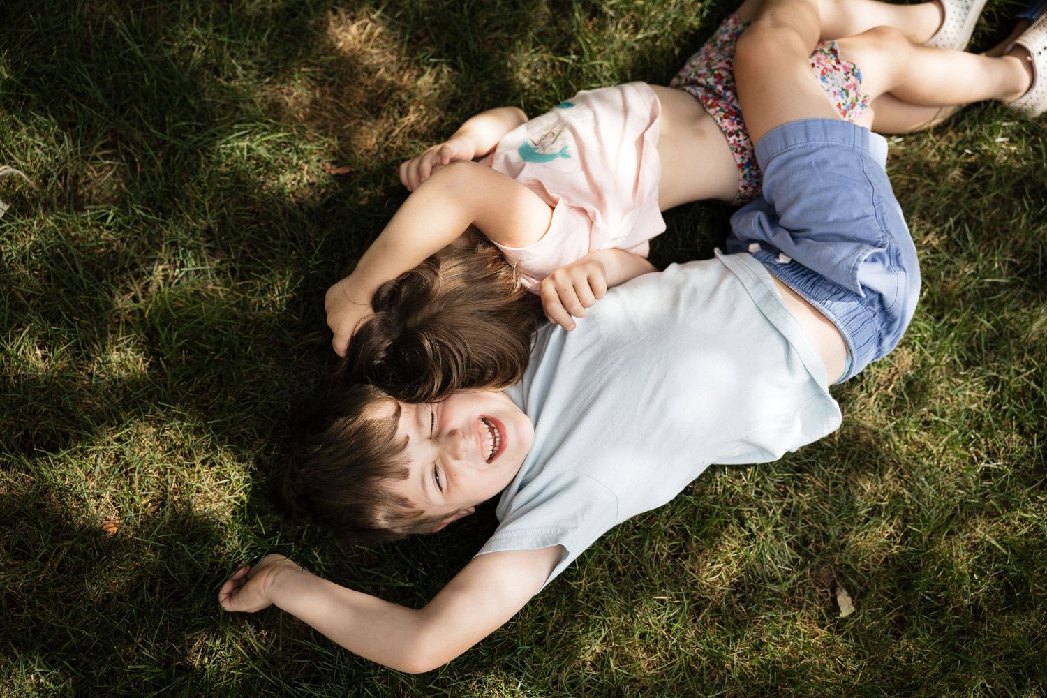 Two children laughing and playing together while lying on green grass outdoors on a sunny day.