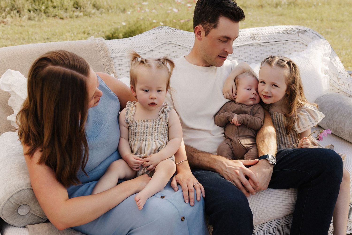 A young family with two toddlers and a baby sits together on an outdoor sofa in a bright, natural setting.