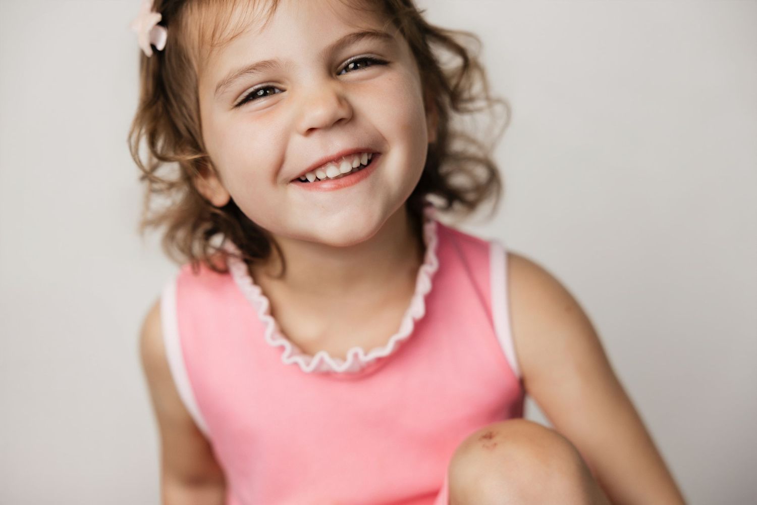 A cheerful young girl in a pink sleeveless top smiles brightly, showcasing her joyful expression against a light background.