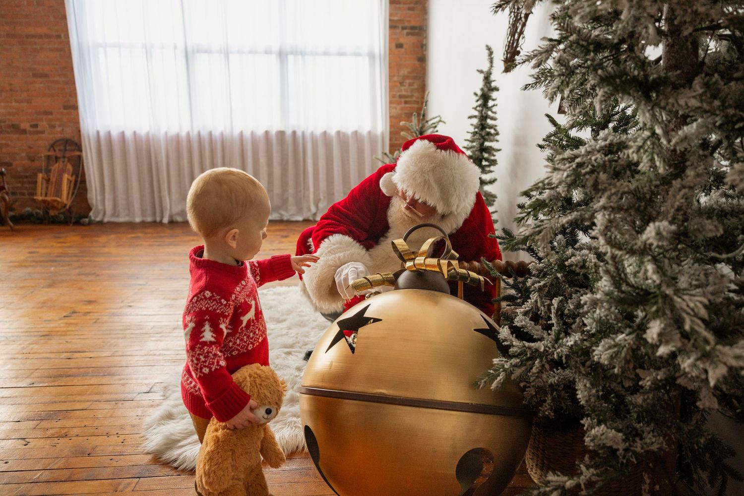 Baby in red outfit touches large golden bell ornament with Santa Claus beside Christmas tree.