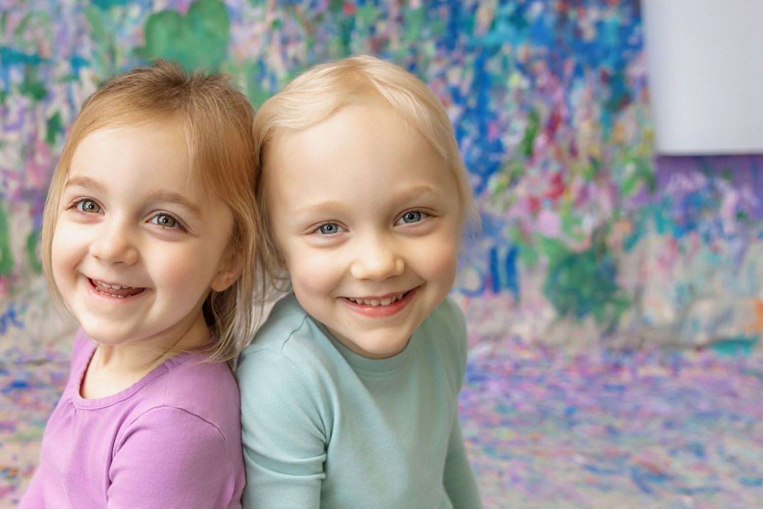 Two smiling young girls sitting back to back in front of a colorful painted background.