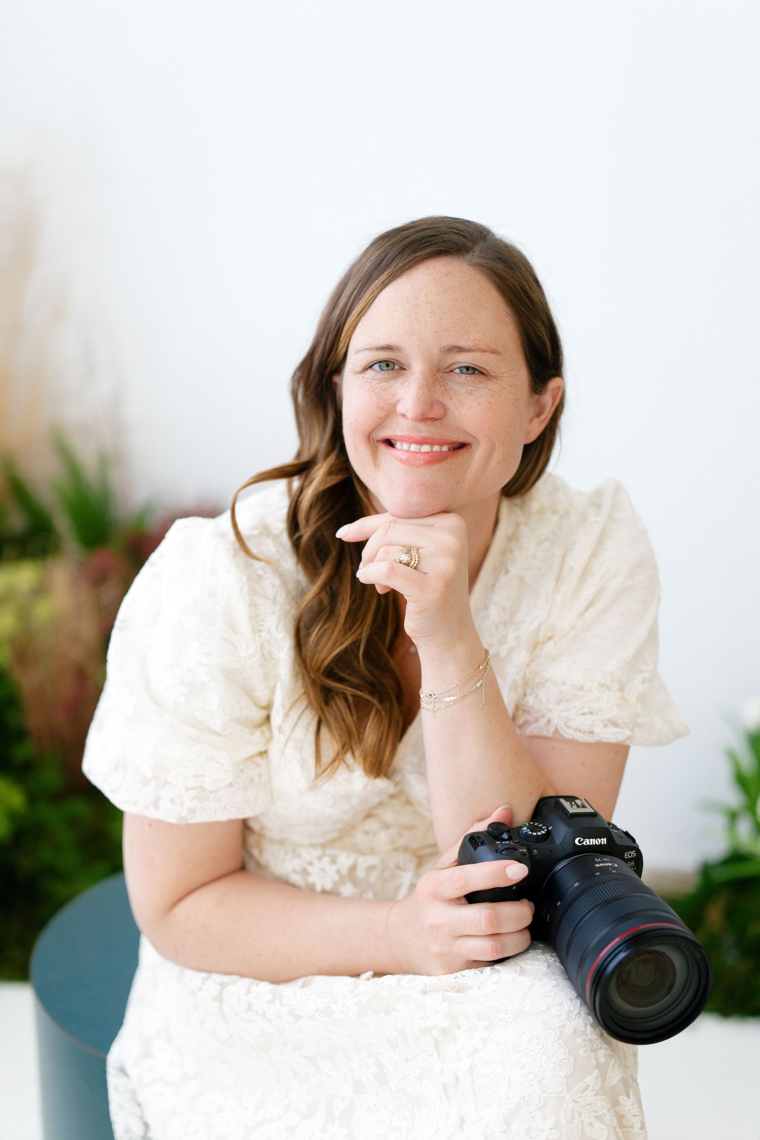 Hilary Mauro in white lace dress posing with Canon camera resting chin on hand.