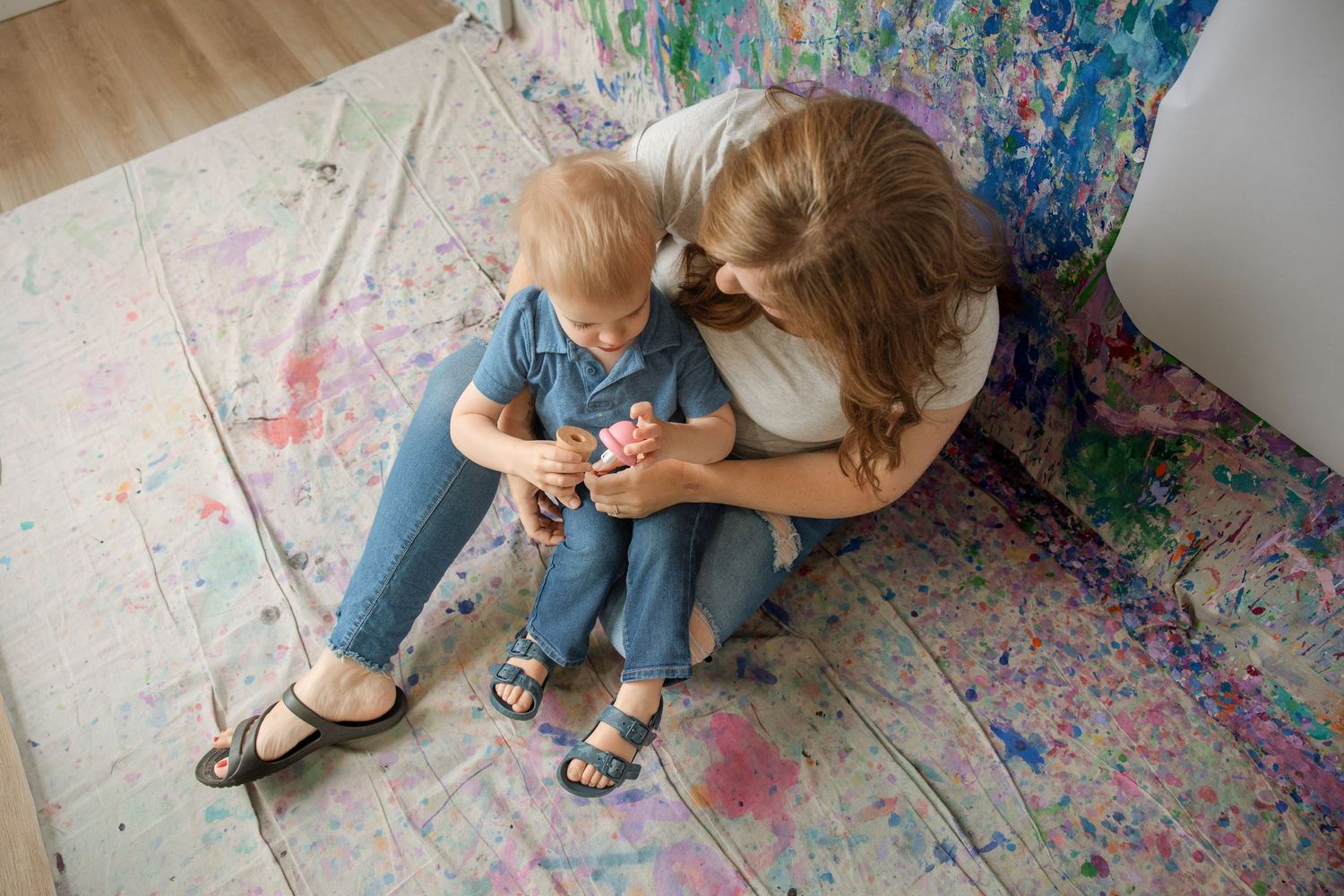 A mother and toddler sit together on a colorful paint-splattered canvas floor in a bright art studio.