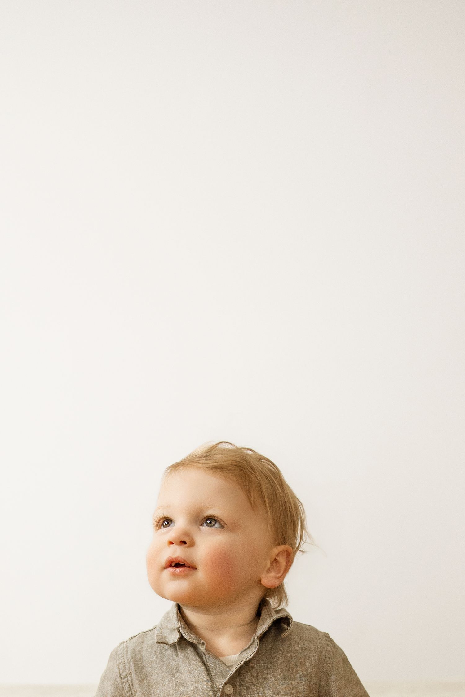 Adorable blonde toddler in gray shirt looking up against a white background.