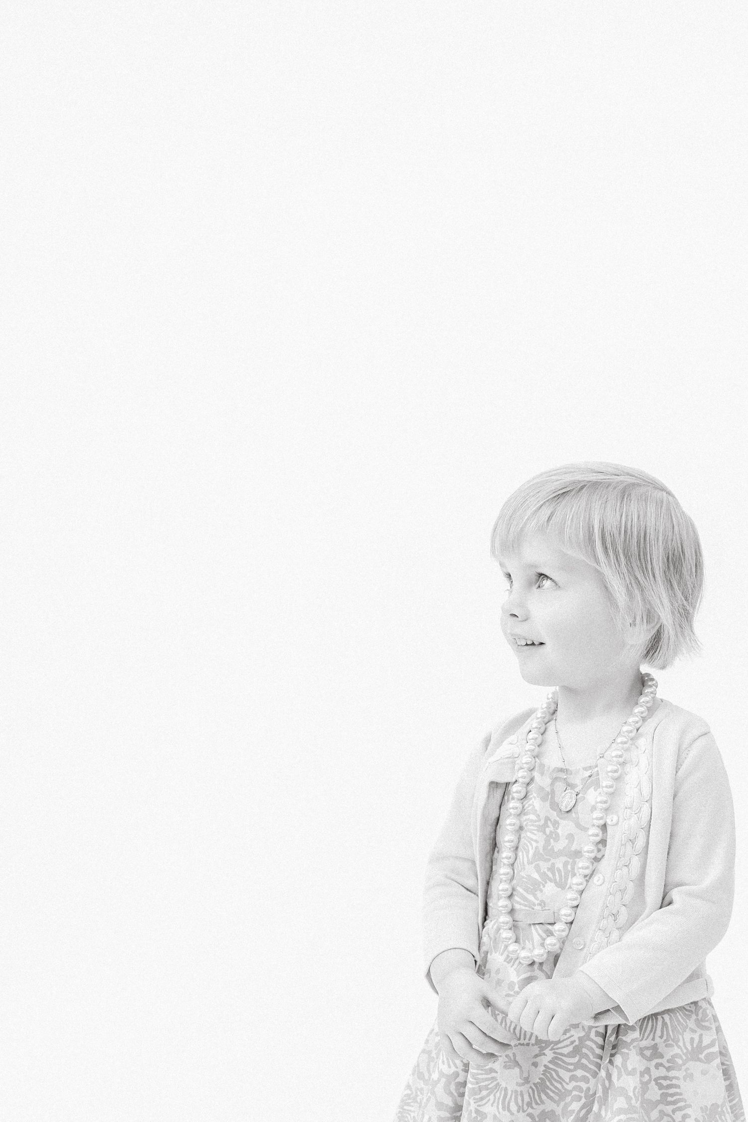 Adorable little girl in floral dress and necklace looking sideways in black and white portrait.