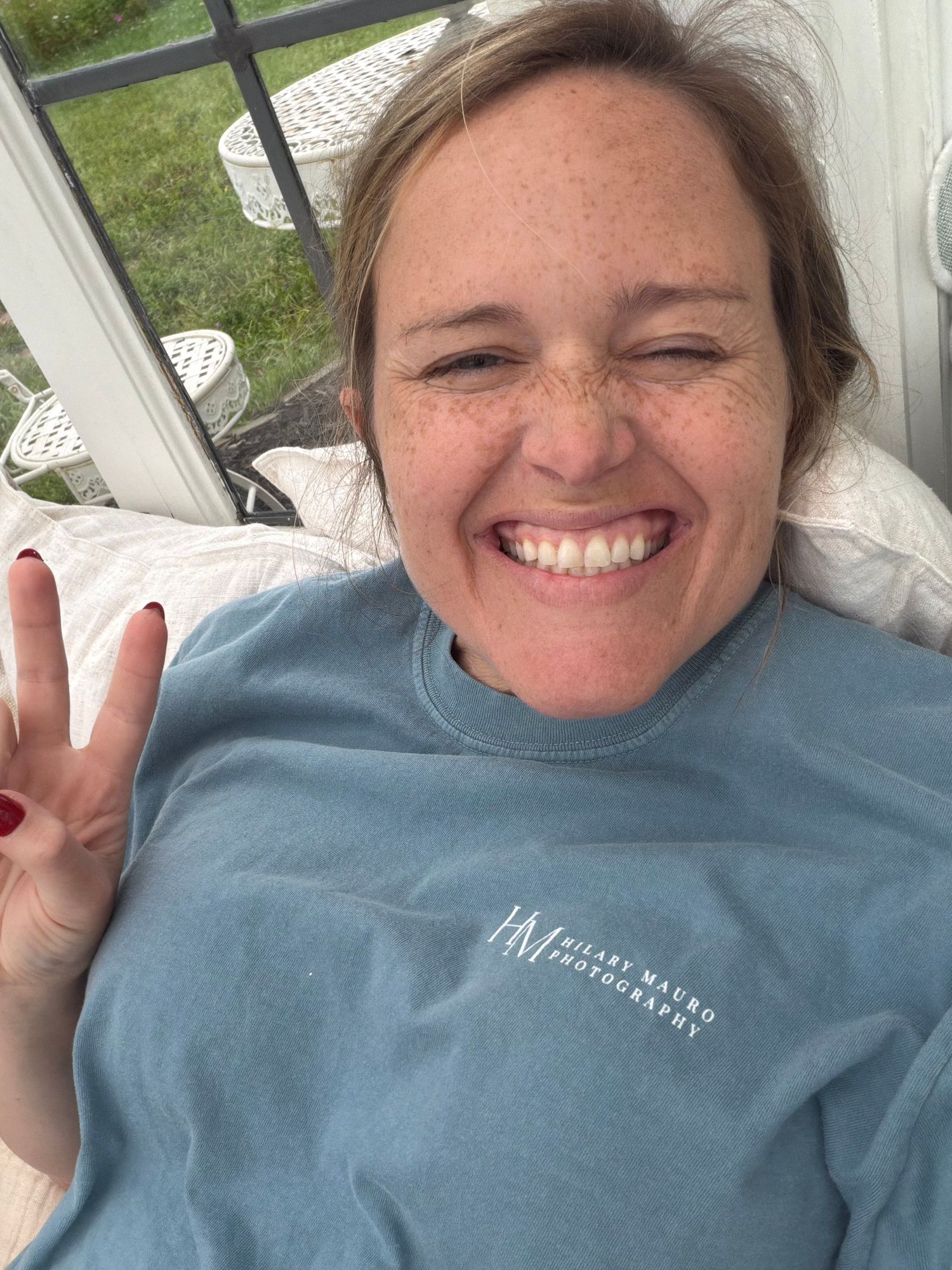 Smiling woman with freckles giving peace sign, wearing light blue HM Hilary Macro Photography shirt, taking selfie outdoors.